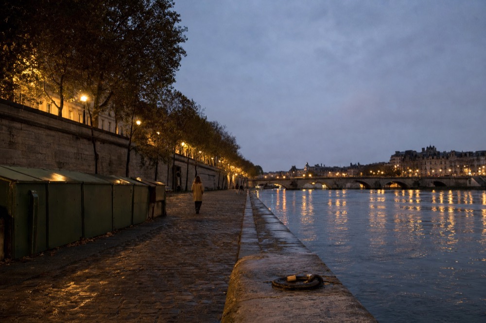 Wet cobbles line a Seine quay at dusk with green riverside boxes, plane trees, lamps, a bridge, and Paris facades across the water.
